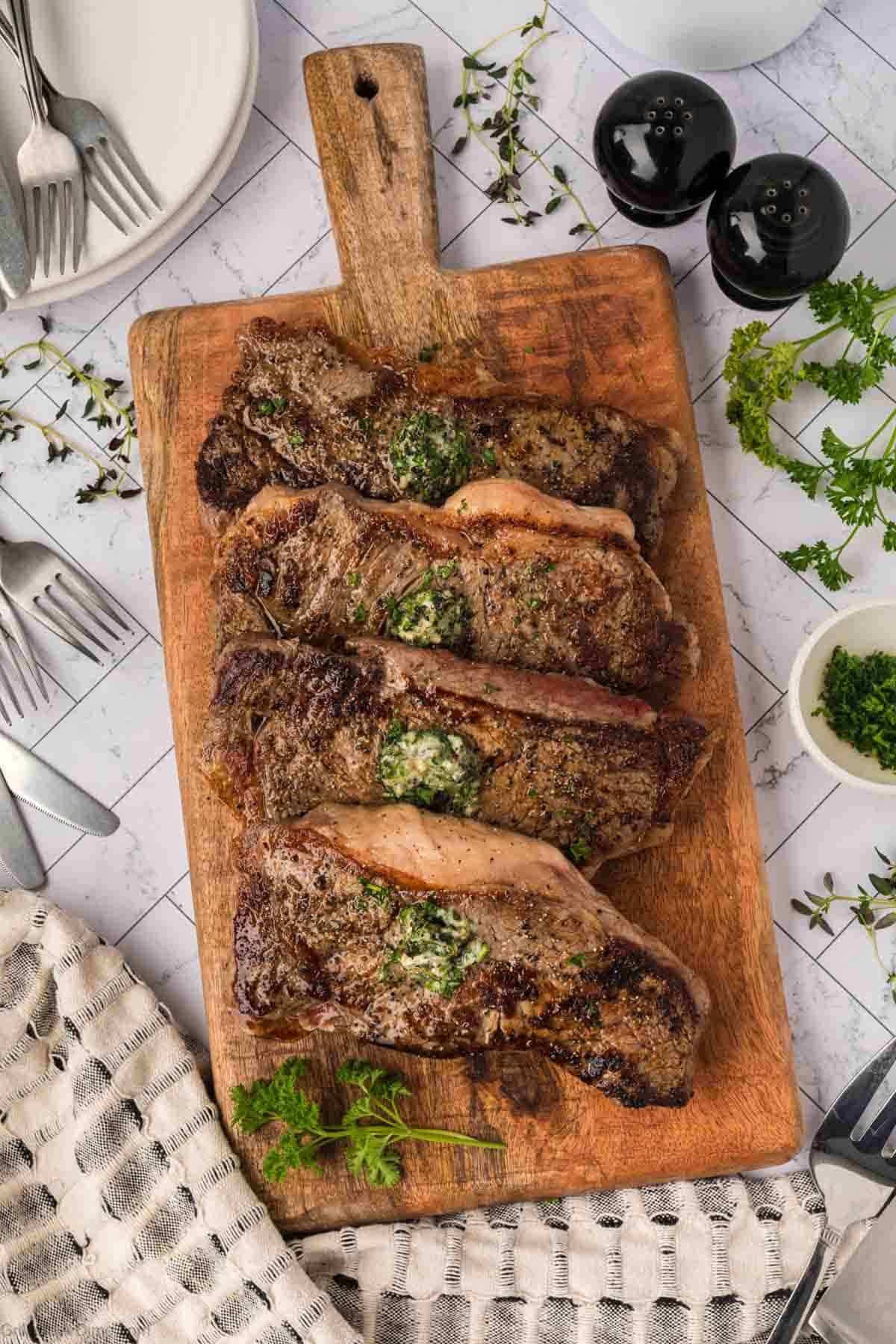 Three cooked New York Strip steaks topped with herb butter and garnished with parsley are arranged on a wooden cutting board, surrounded by plates, cutlery, pepper shakers, and a small bowl of herbs on a tiled surface—perfect for the Blackstone grill.