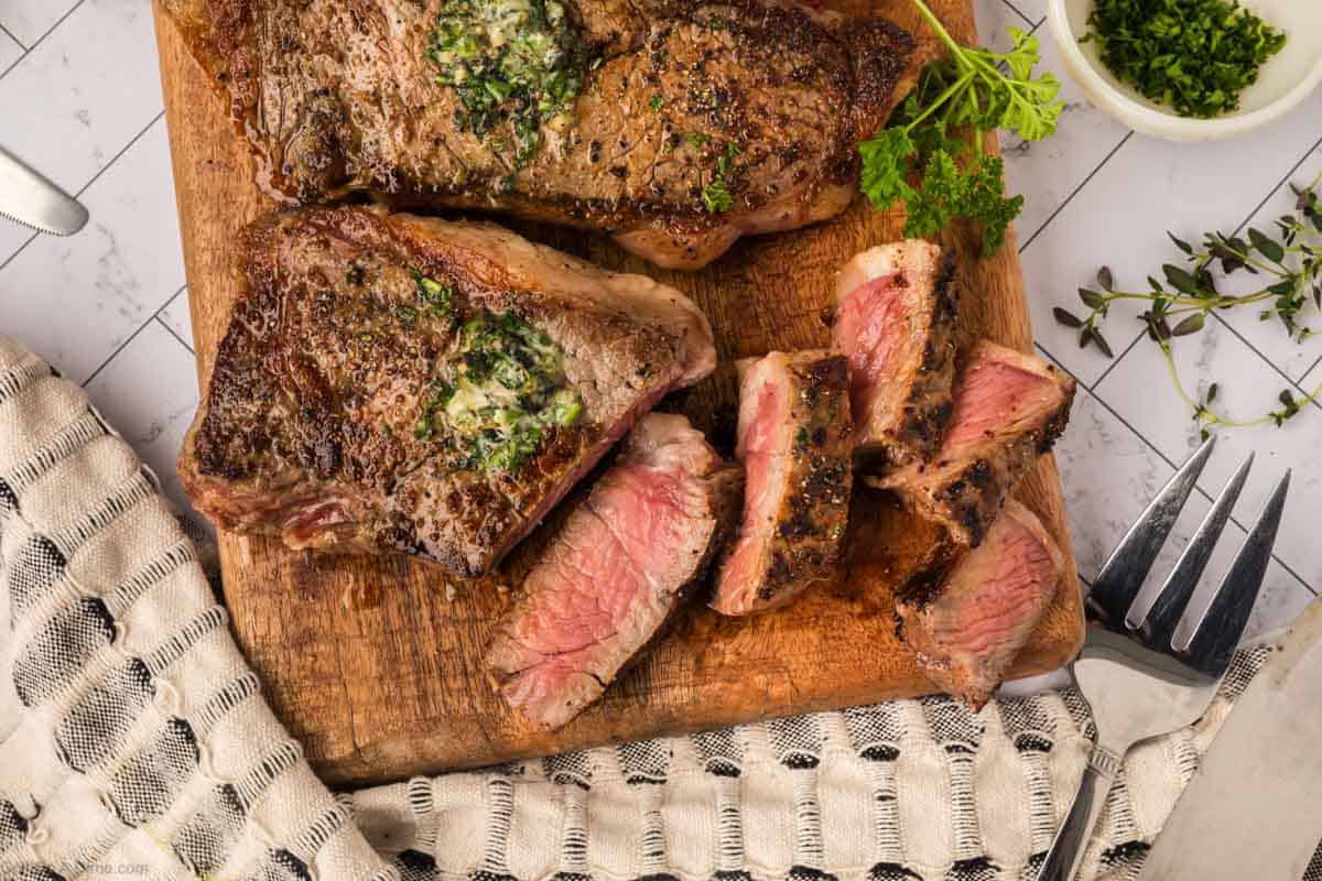 A cooked New York Strip steak topped with herb butter sits on a wooden board. Next to it are sliced pieces, showing a pink, medium-rare center. A fork, herbs, and a cloth napkin are nearby—a delicious result from the Blackstone grill.