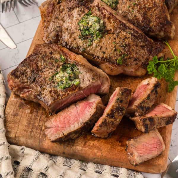 Sliced grilled New York Strip steak served on a wooden board, topped with herb butter. The meat is cooked medium rare, showing a pink center. Fresh parsley is placed next to the steak, with a fork and napkin visible nearby.