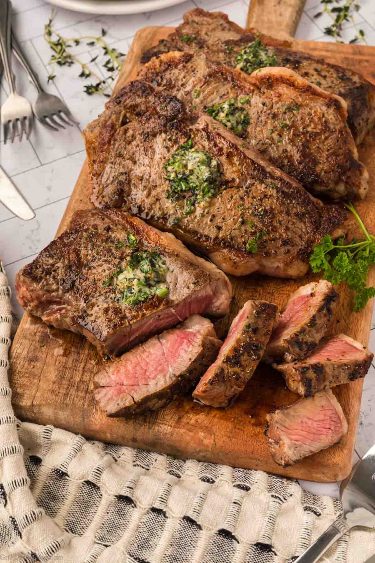 Juicy New York Strip steaks, grilled on a Blackstone and topped with herb butter, are arranged on a wooden cutting board. Some slices reveal a medium-rare center, with fresh herbs and cutlery visible in the background.