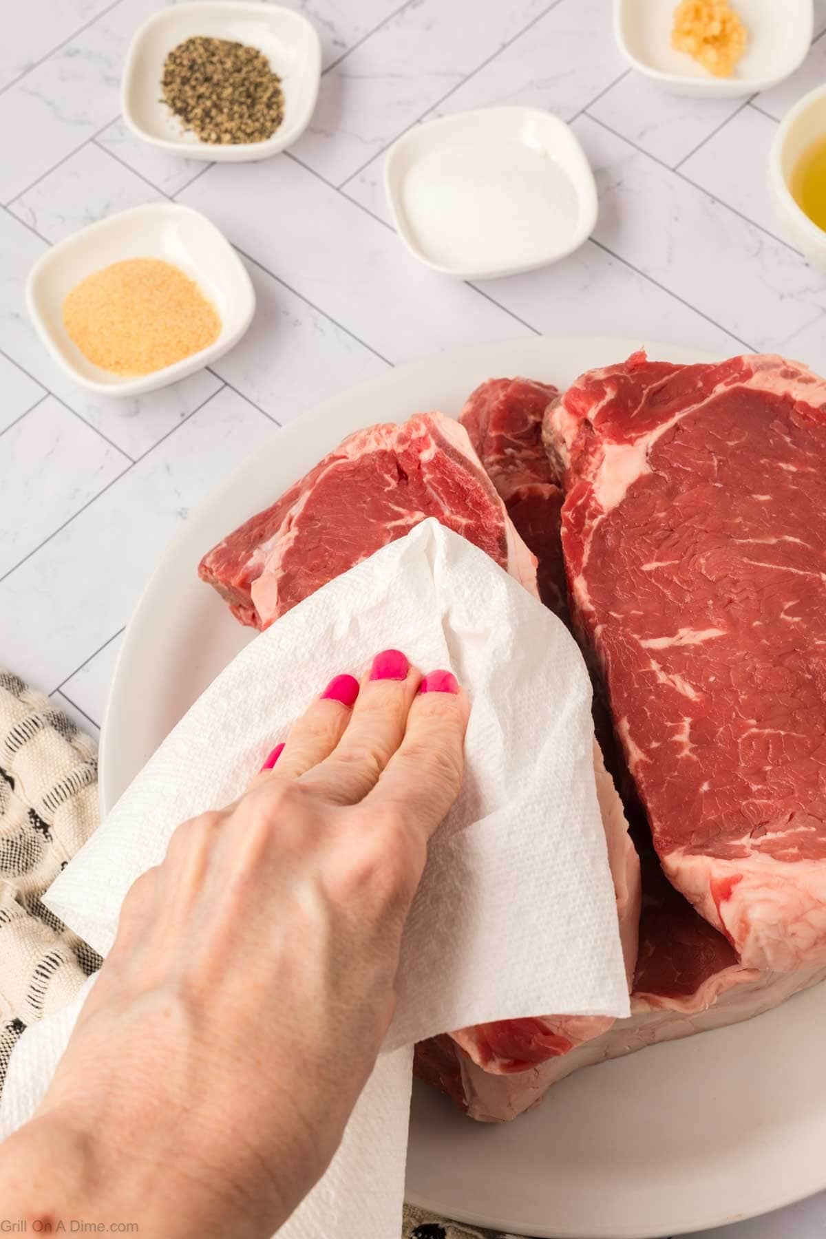 A hand with pink fingernails uses a paper towel to pat dry raw New York Strip steaks on a white plate. Small bowls with seasonings like salt, pepper, garlic, and oil are visible in the background on a tiled surface.
