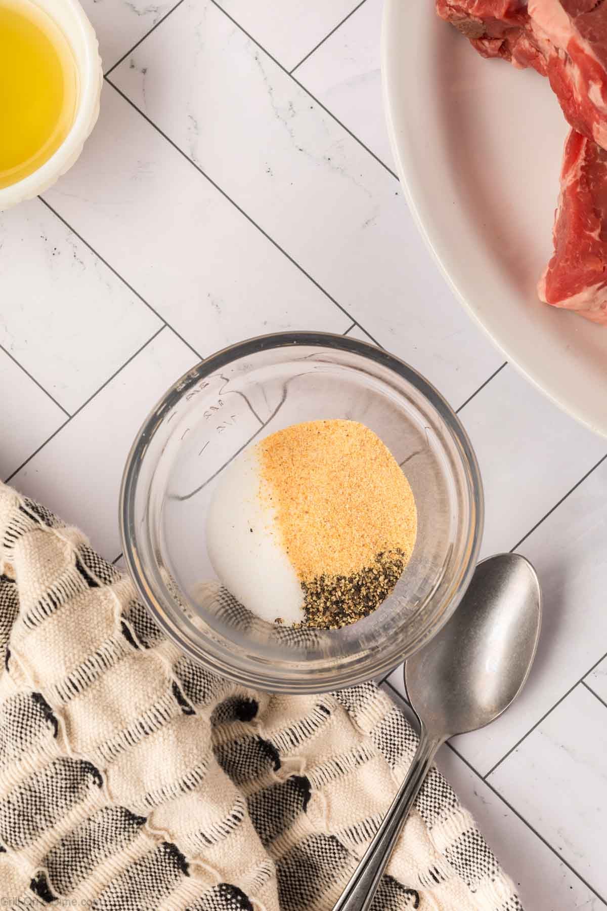 A small glass bowl with salt, pepper, and garlic powder sits on a checkered cloth next to a metal spoon. Nearby, there is a plate with raw New York Strip steak and a cup of oil on a white tiled counter.