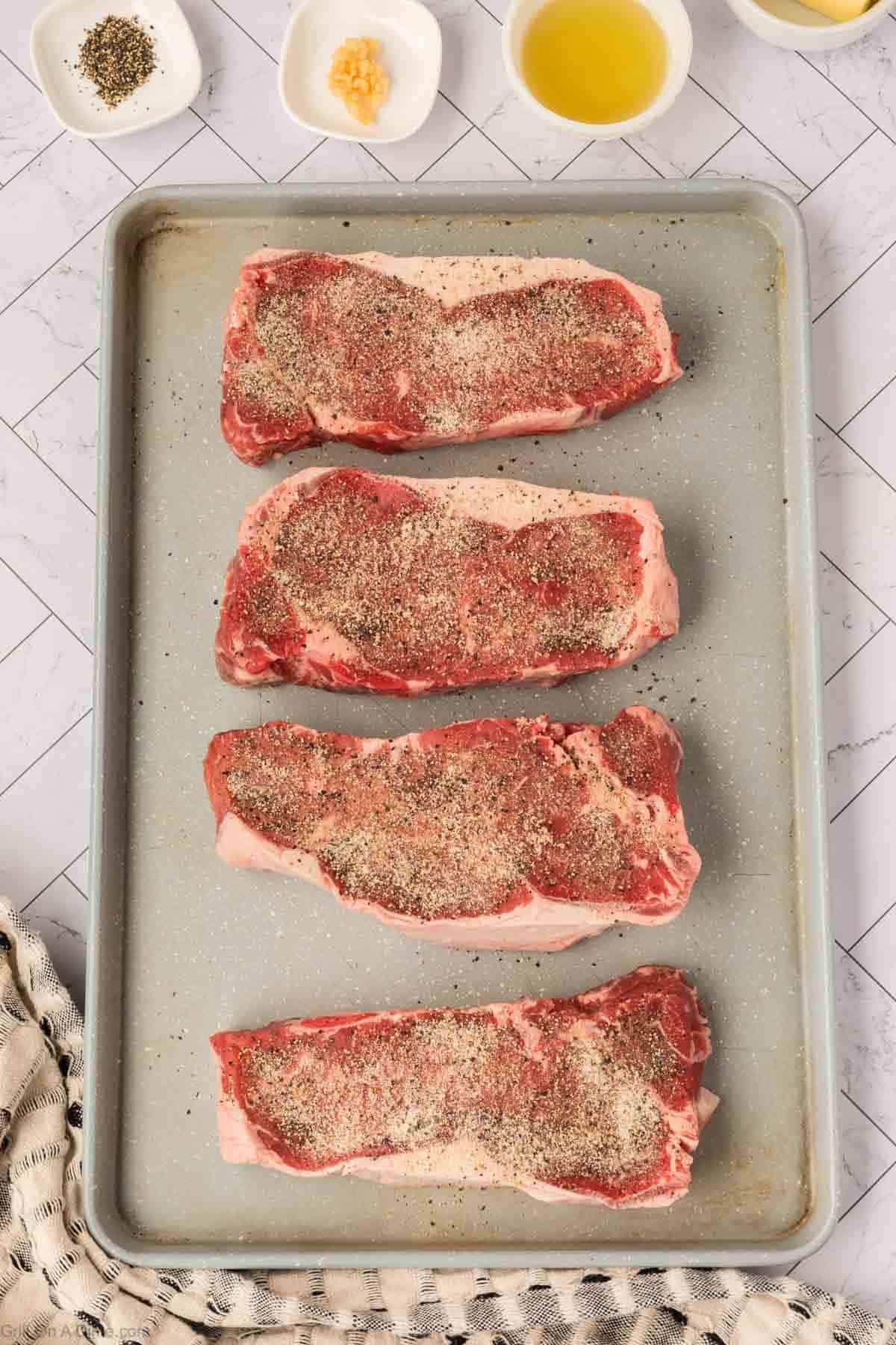 Four raw, seasoned New York Strip steaks are arranged in a row on a baking sheet. Small bowls with olive oil, minced garlic, and spices sit at the top, and a striped towel is in the lower corner.