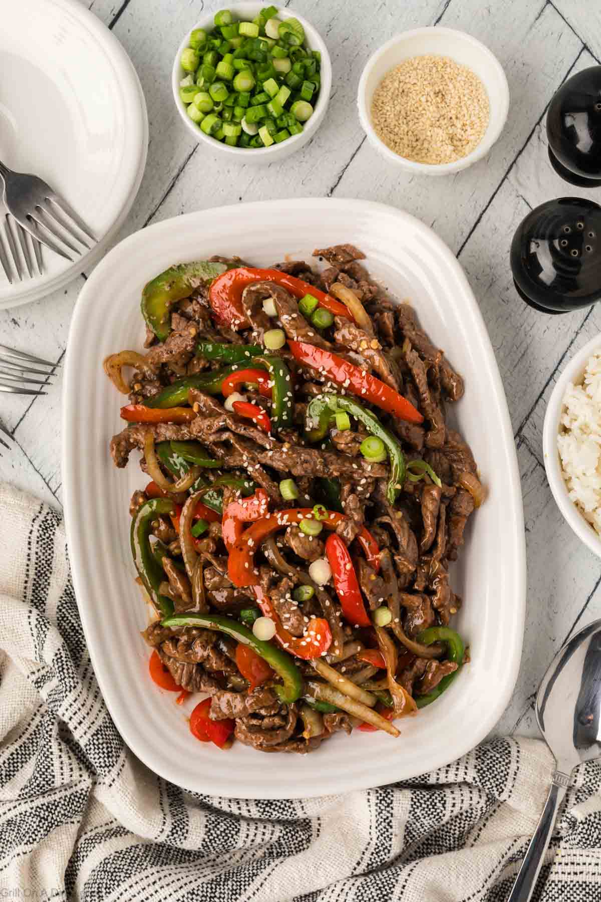A platter of stir-fried beef with red and green bell peppers and onions is garnished with sliced green onions and sesame seeds.