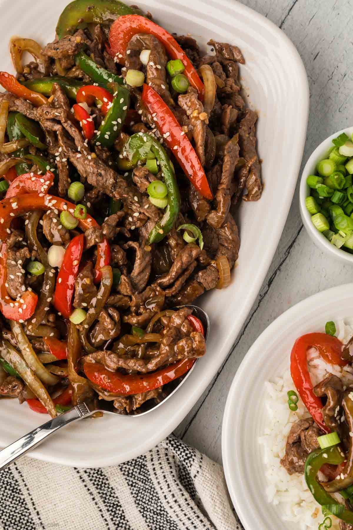 A platter of stir-fried beef with red and green bell peppers, onions, and sesame seeds is garnished with chopped green onions.