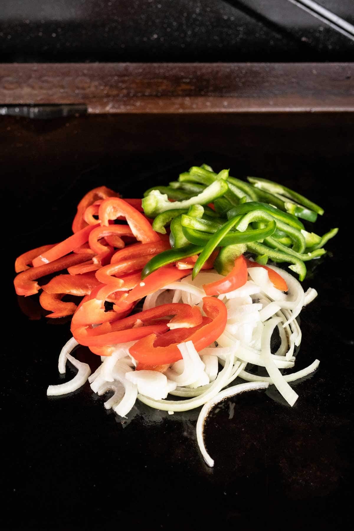 Sliced red and green bell peppers with white onions are arranged in small piles on a Blackstone griddle.