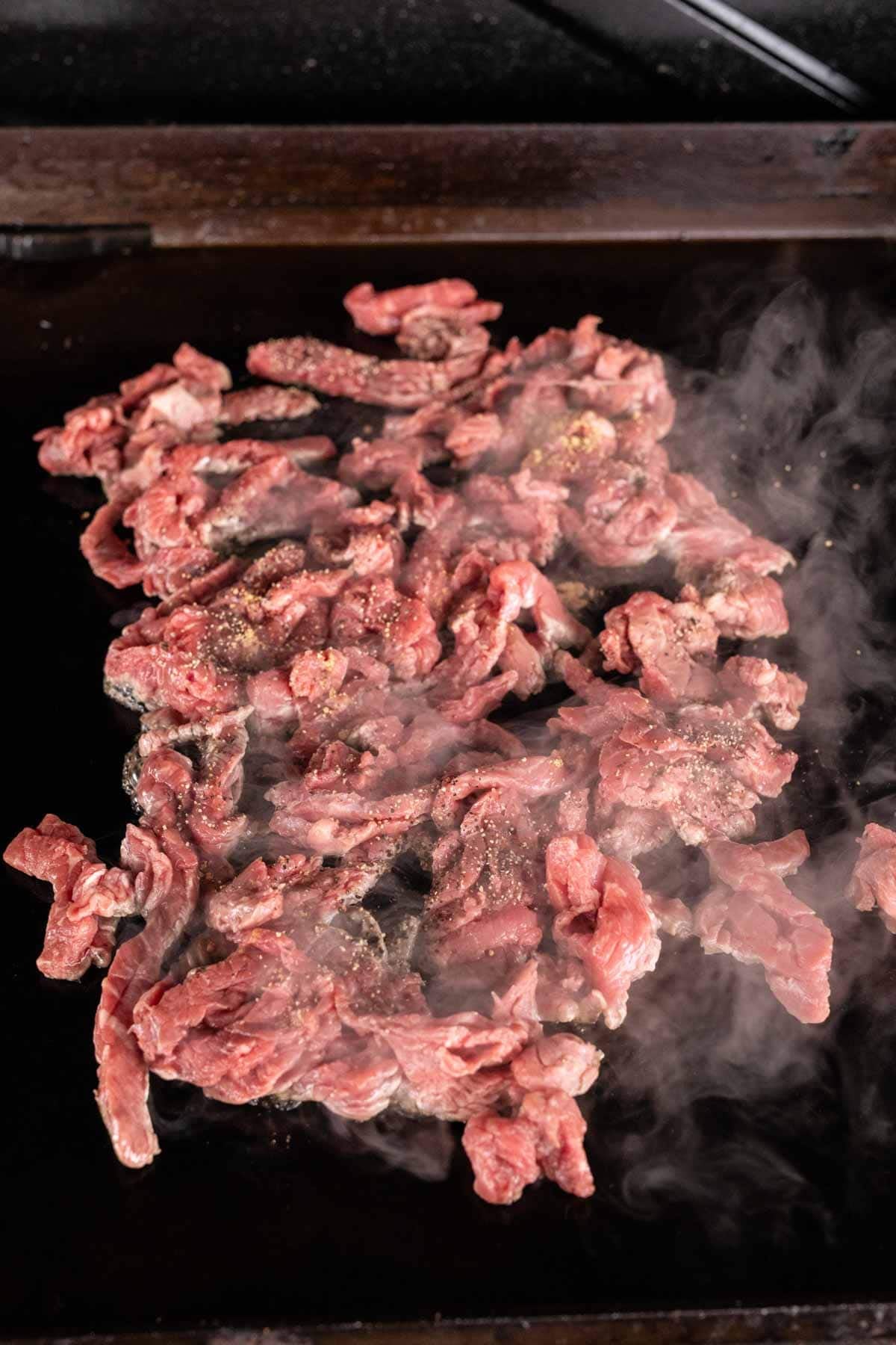 Thin slices of raw beef are sizzling and beginning to cook on a hot Blackstone griddle, with steam rising from the meat.