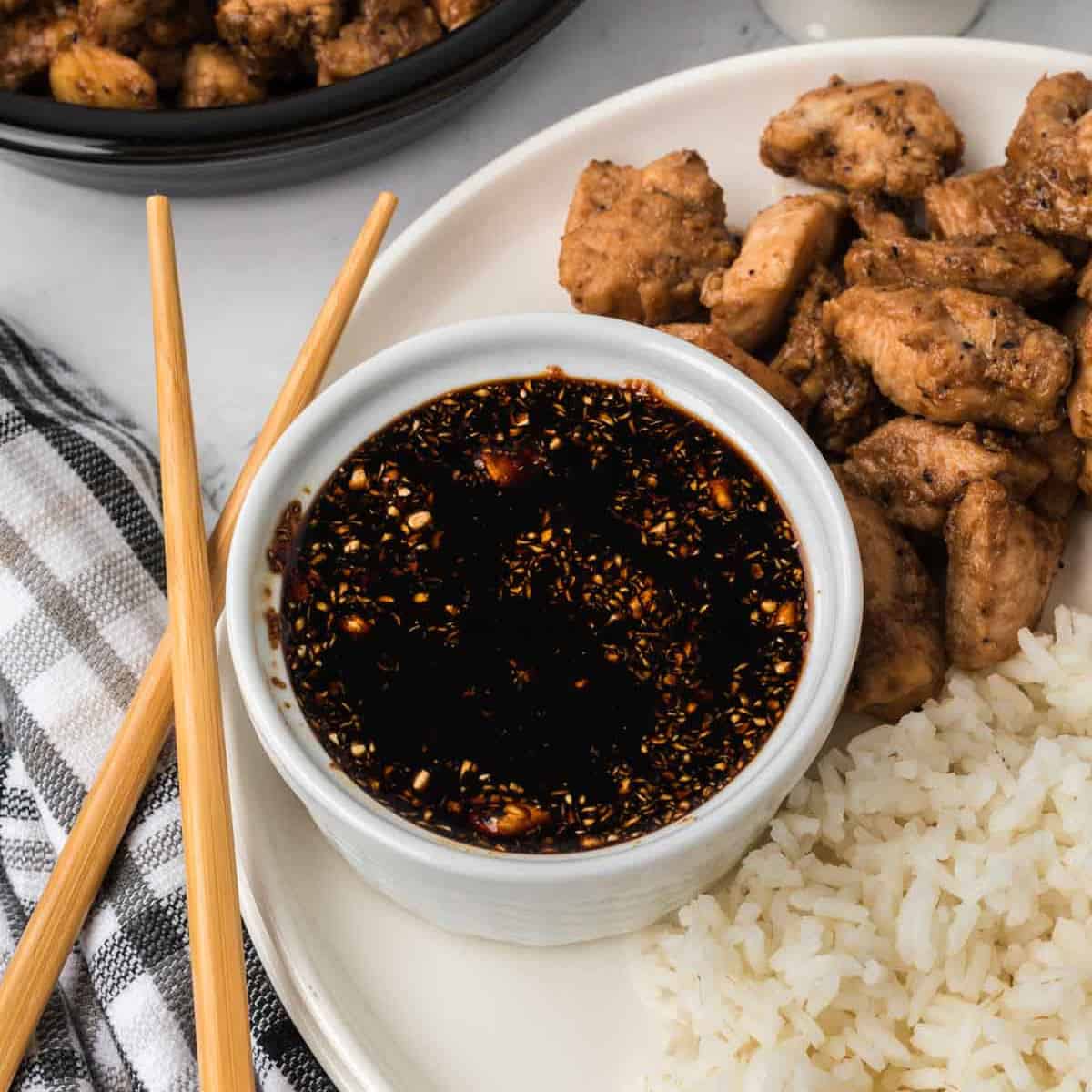 A bowl of dark Hibachi Sauce sits on a plate with white rice and cooked chicken pieces. Wooden chopsticks rest beside the bowl, and a checkered cloth is nearby.