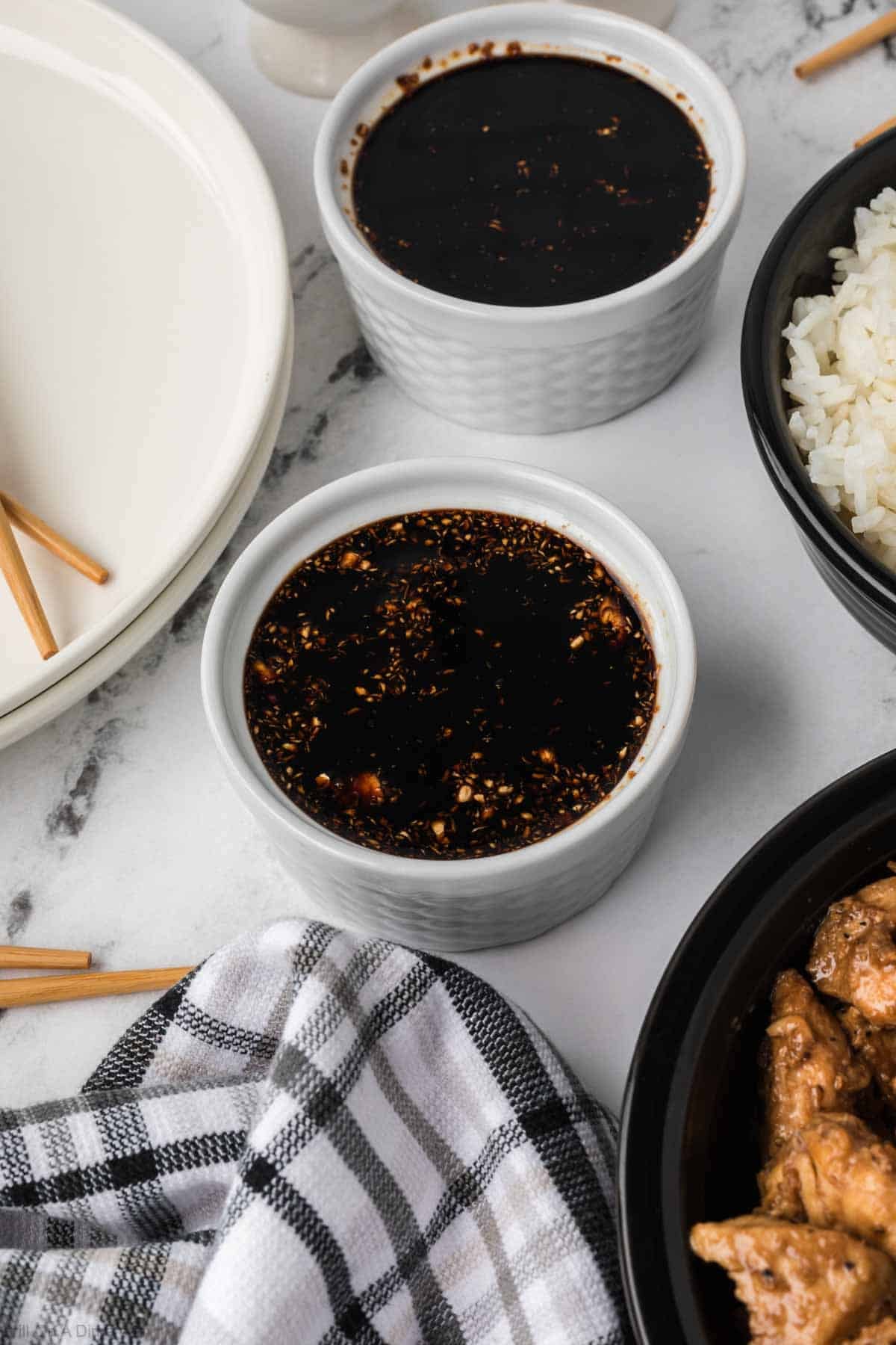 Small white bowl of dark sauce with a side of bowl of white rice and cooked chicken. 