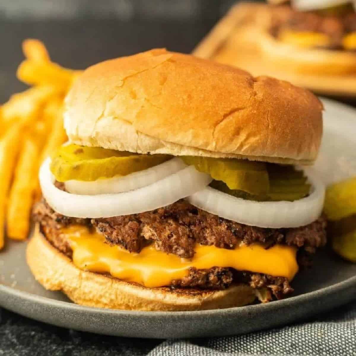 A cheeseburger with melted cheese, sliced onions, pickles, and a beef patty in a bun, served with French fries on a plate.