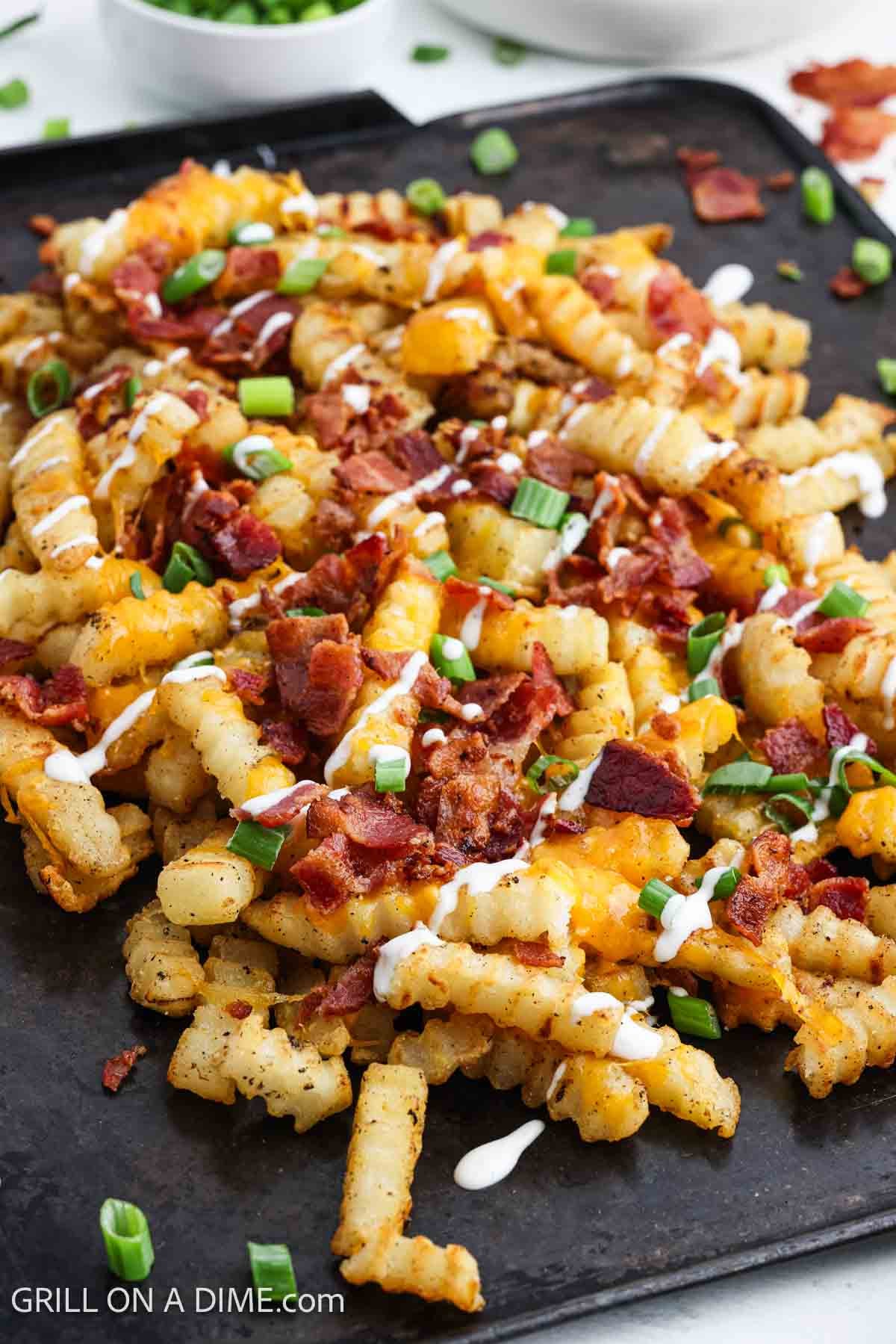 Cheese fries topped with melted cheese, chopped bacon, green onions and a drizzle of ranch on a black serving platter. 