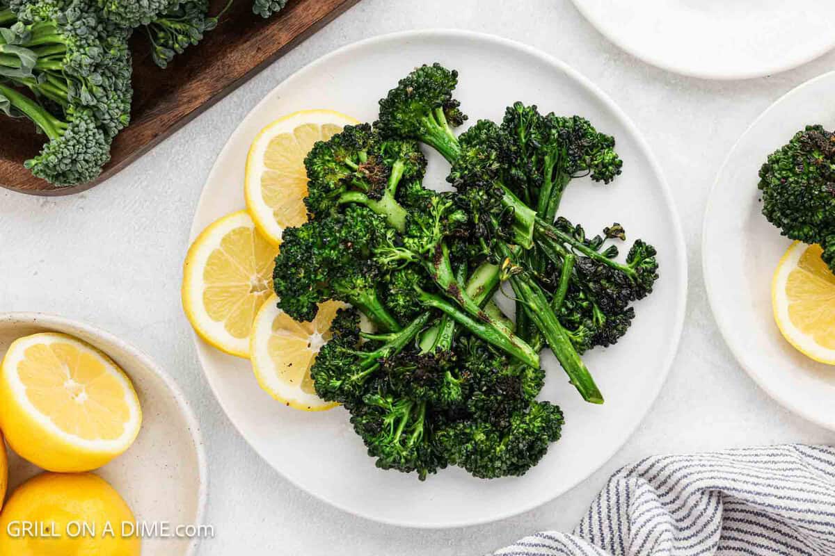 A plate of grilled broccolini arranged with lemon slices on a white plate, surrounded by fresh lemons and a striped napkin.