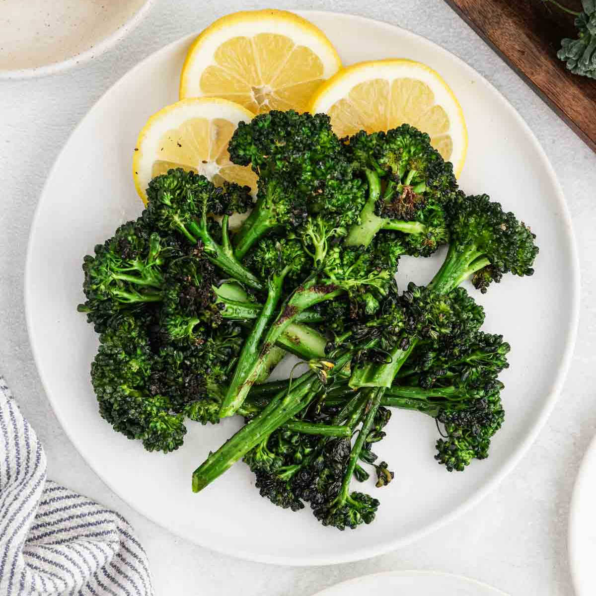 A white plate with Grilled Broccolini garnished with three lemon slices, viewed from above.