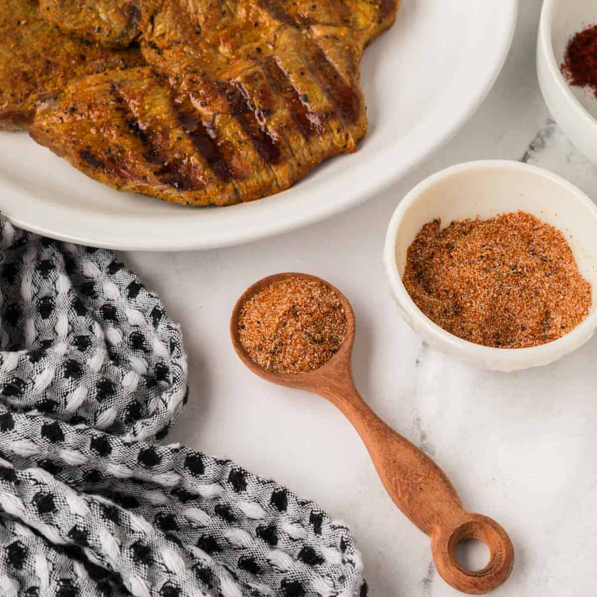 Grilled meat on a white plate, paired with a wooden spoon filled with Outback Steak Seasoning with a small bowl of seasoning on the side.