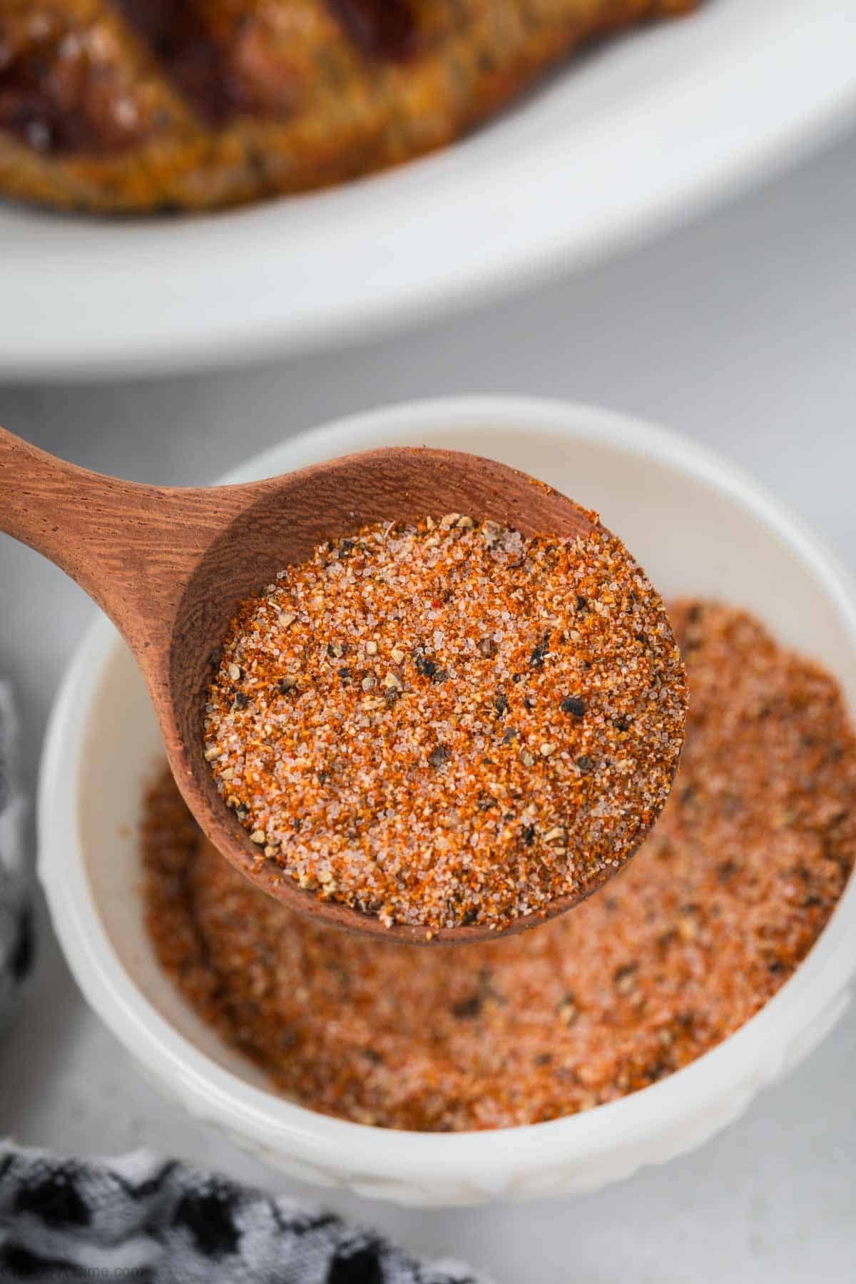 A wooden spoon holds a blend of Seasoning over a small white bowl filled with the same spice mix. 