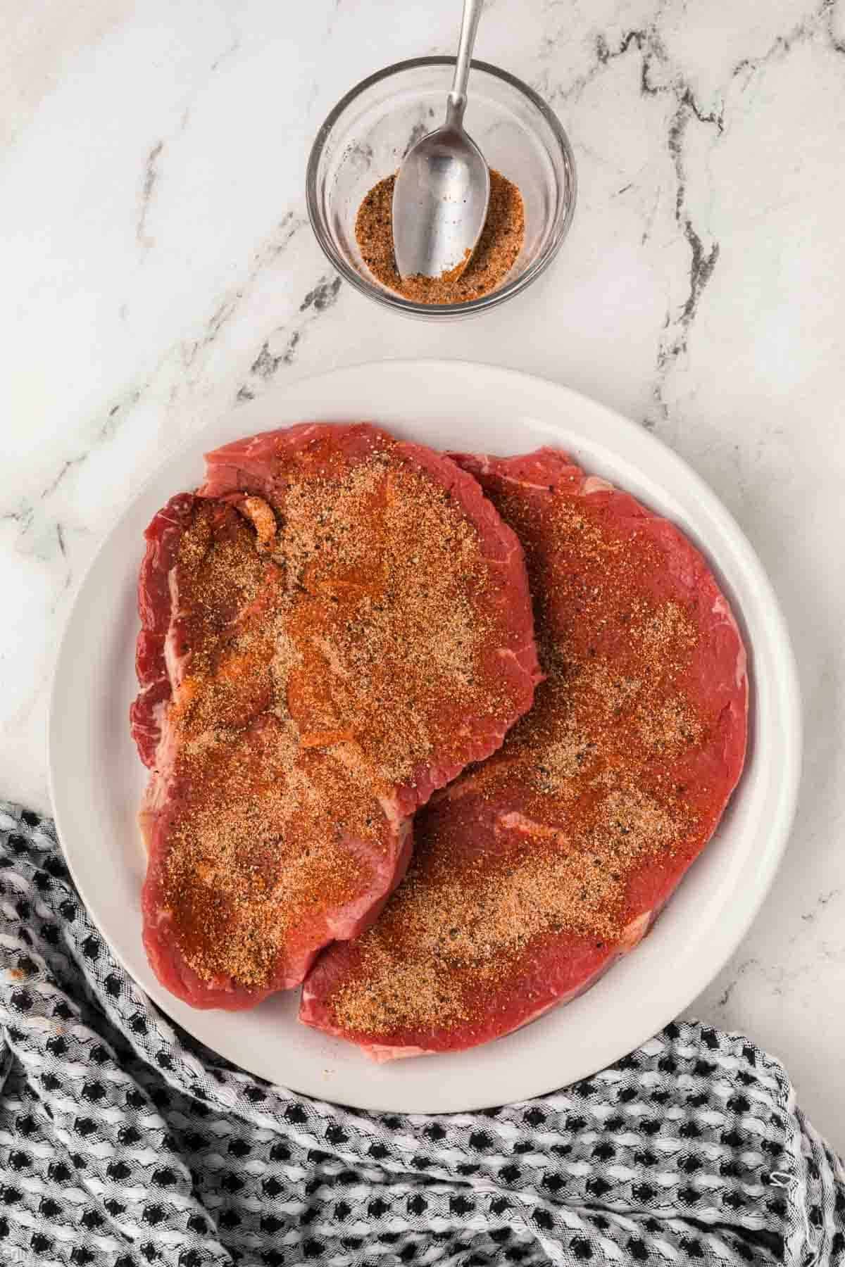 Two raw steaks coated with seasoning sit on a white plate beside a small glass bowl of extra seasoning with a spoon placed on top. 