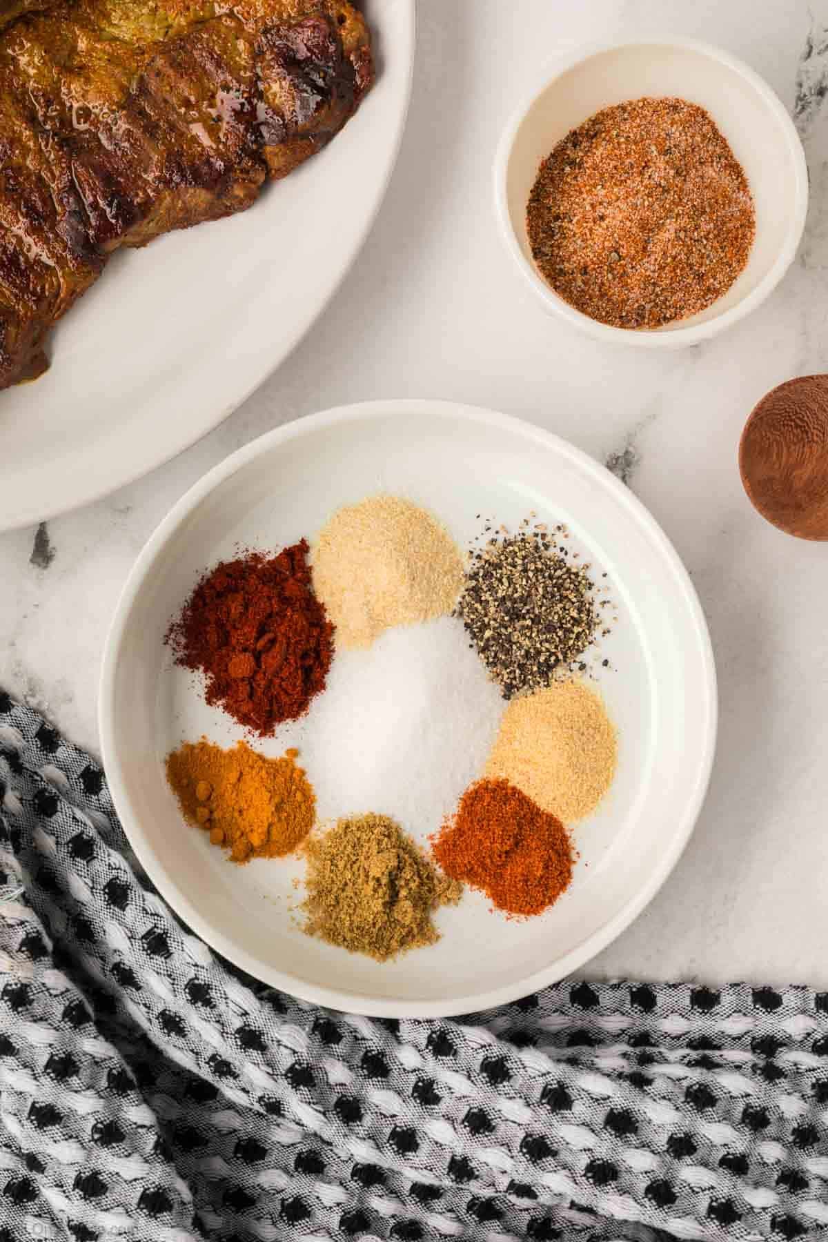 A white plate displays small piles of various spices with a small bowl of seasoning blend and plate of cooked steak on the side. 