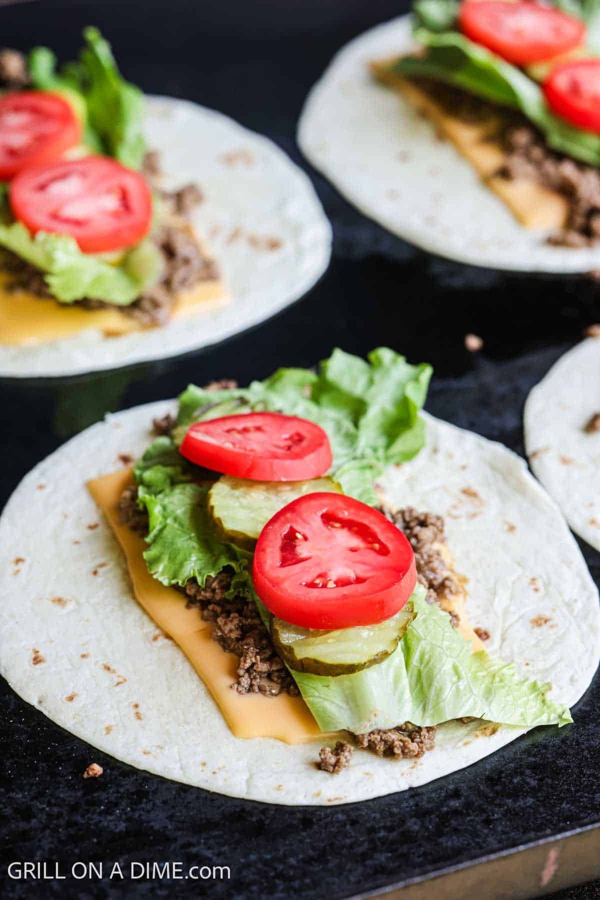 Tortilla on the flat top griddle topped with slice cheese, cooked ground beef, lettuce and tomatoes.