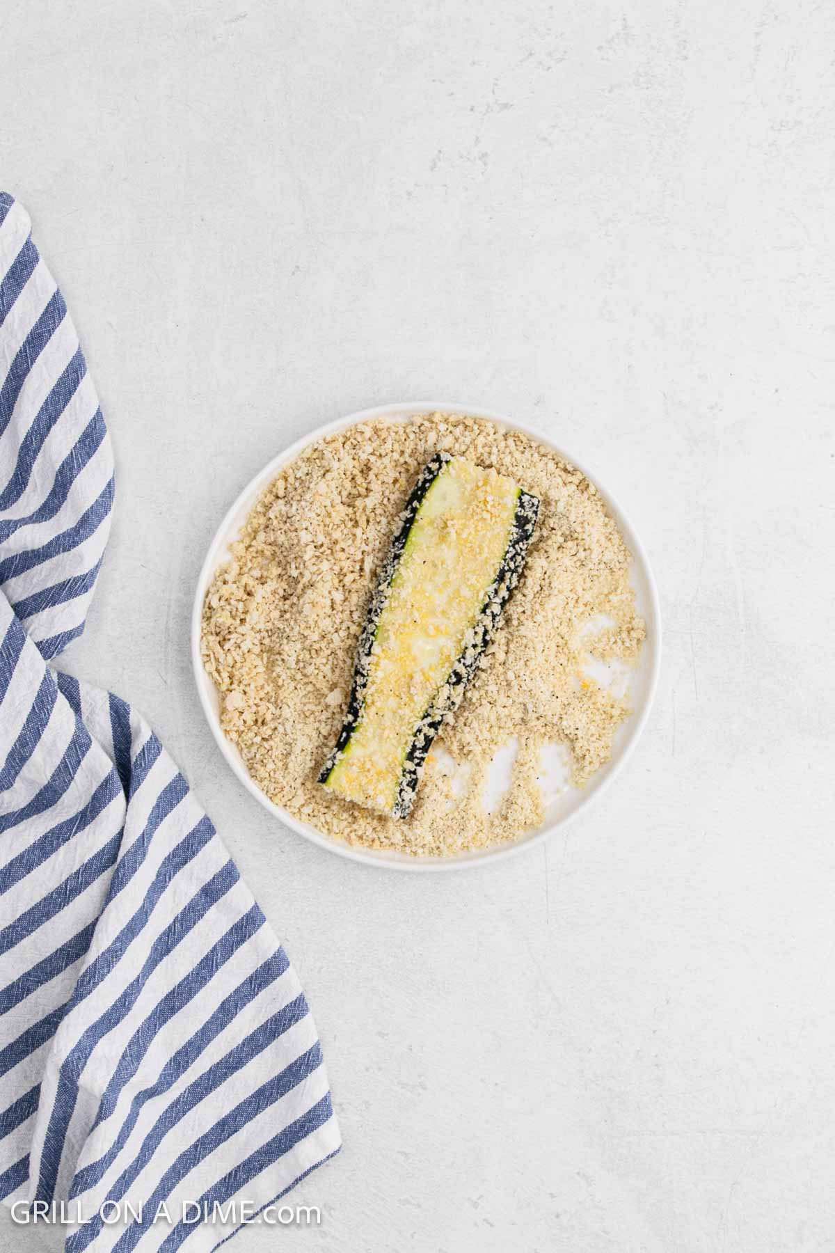 Dipping the slice zucchini in the parmesan mixture on a plate.