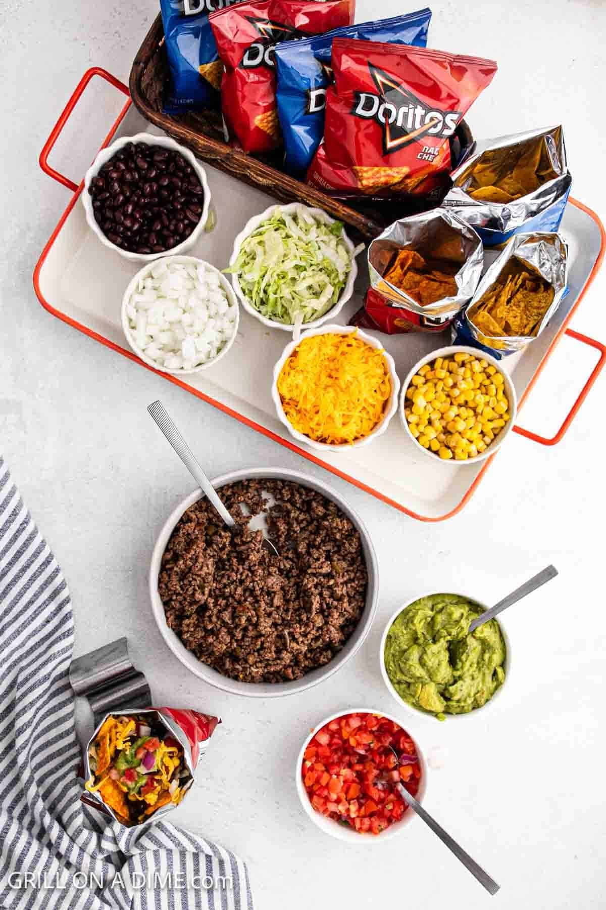 Small bowls of black beans, onions, lettuce, cheese, corn, ground beef, guacamole, and salsa with small bags of Doritos on the side.