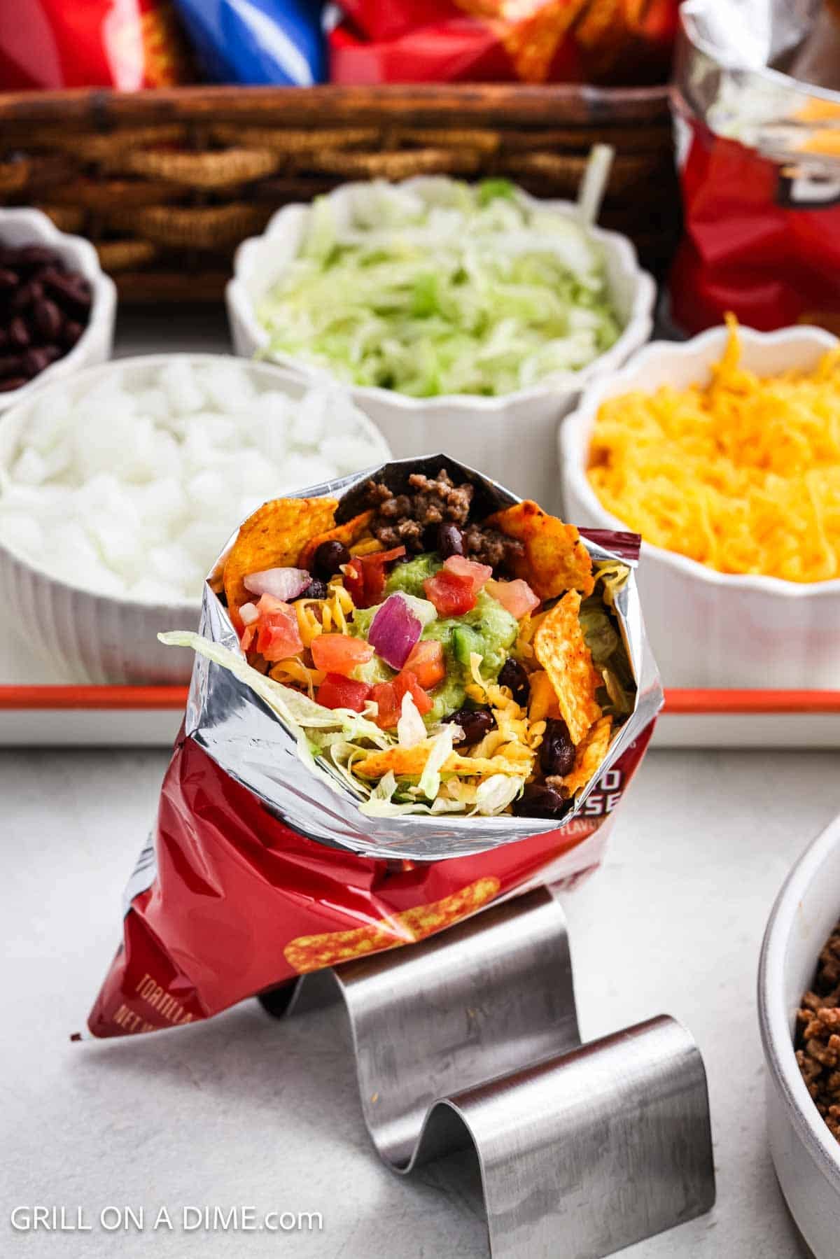 A bag of Doritos chips filled with beef, lettuce, cheese, pico and guacamole with small bowls of toppings in the background.